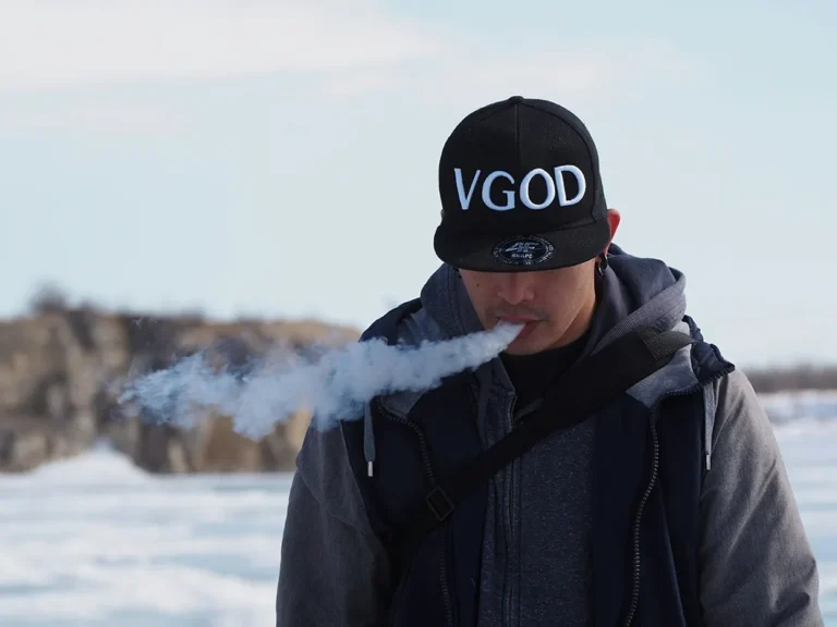 一位戴著「VGOD」品牌帽子和灰色連帽衫的成年男性老玩家，在模糊的雪地戶外冬日場景中專注地吞吐厚實的白色電子煙霧化蒸汽。這是一張用於現代替煙方案指南的特色圖片。