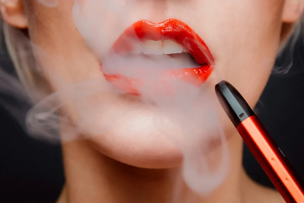 一幅女性特寫圖片，塗有鮮紅唇膏的嘴唇正呼出電子煙的白色霧化蒸汽，手中握著一支時尚的深橙紅色電子煙主機。此圖展示了現代替煙方案的使用場景。